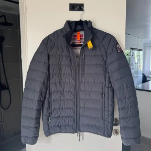 Svart dunjacka från Parajumpers - Snygg svart dunjacka från Parajumpers med klassisk patch på ärmen och gul detalj vid kragen. Jackan har quiltad design, dragkedjor på fickorna och hög krage. Perfekt för kyliga dagar och riktigt stilren look.