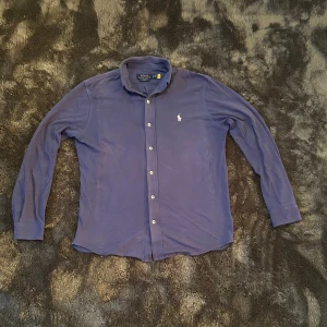 Blå långärmad skjorta från Polo Ralph Lauren - Säljer en blå långärmad skjorta från Polo Ralph Lauren med klassisk krage och knappar framtill. Skjortan har det ikoniska broderade logotypen på bröstet och är tillverkad i lätt bomullsmaterial. Perfekt för en clean och stilren look.