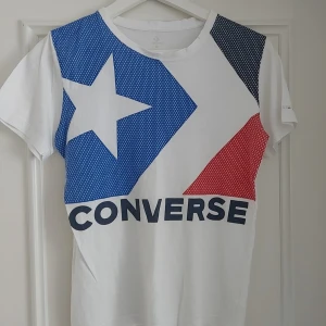 Vit Converse t-shirt med grafiskt tryck 10-12år - Snygg vit t-shirt från Converse med stort grafiskt tryck i blått, rött och svart framtill. T-shirten har korta ärmar, rund halsringning och är gjord i mjuk bomull. Perfekt för dig som gillar streetstyle och vill sticka ut med en ikonisk logga.