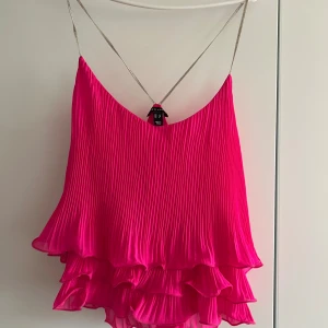 Plisserad rosa linnetopp med volanger - Supersnygg plisserad linnetopp från Zara i starkt rosa med tunna silvriga axelband. Toppen har volangkant och dubbla lager nertill för extra flowig känsla. Perfekt för dig som vill sticka ut med färg och coola detaljer.