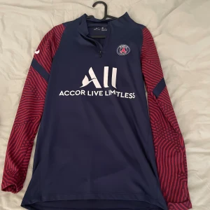PSG långärmad träningströja Nike - Paris Saint-Germain långärmad träningströja från Nike i mörkblått med röda ärmar och unikt mönster. PSG-logga på bröstet och Accor Live Limitless-tryck på framsidan. Materialet är lätt och andas, perfekt för fotbollsträning. 