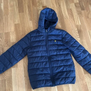 Blå pufferjacka från Lyle & Scott - Snygg marinblå pufferjacka från Lyle & Scott med huva och dragkedja framtill. Jackan har det klassiska gula logomärket på bröstet och är quiltad för extra värme. Perfekt för kyliga dagar och enkel att matcha med olika outfits.