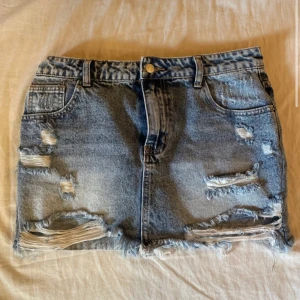 Blå jeanskjol med slitningar - Säljer en blå jeanskjol. Kjolen har klassisk femficksdesign, bälteshällor och knäppning fram. 