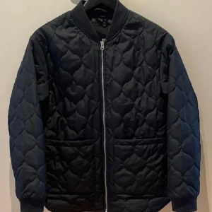 Svart quiltad bomberjacka - Svart quiltad bomberjacka med dragkedja framtill och två stora fickor. Jackan har ribbade muddar vid ärmslut och hals, samt ett snyggt mönster i tyget. Perfekt för dig som gillar en stilren och modern look.