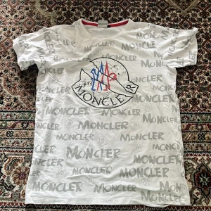 Vit Moncler t-shirt med logotyptryck - Vit t-shirt från Moncler med grått all-over logotyptryck och stor logga framtill i blått och rött. Klassisk rund halsringning med röd kant. Perfekt för dig som gillar streetwear och vill sticka ut med en ikonisk design.