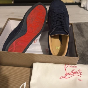 Mörkblå sneakers från Christian Louboutin - Snygga mörkblå sneakers från Christian Louboutin i mocka med klassisk röd sula. Skorna har rund tå, snörning och diskret broderad logga på sidan. Insidan är ljusbeige och sulan är platt. Kommer med originalkartong och dustbag.