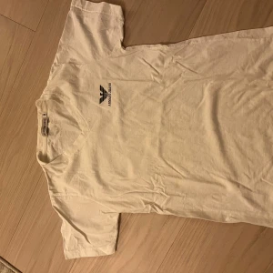 Vit t-shirt från Armani Jeans - Vit t-shirt från Armani Jeans med svart logga och text på bröstet. Klassisk rund halsringning och korta ärmar. Tillverkad i mjuk bomull som känns skön mot huden. Perfekt för dig som gillar stilrena och enkla plagg med exklusiv känsla.