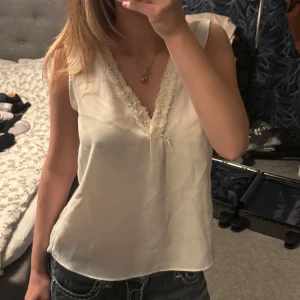 Vit pärlprydd blus från Zara - Supersöt vit blus från Zara Basic i storlek XS. Toppen är ärmlös med v-ringning och har dekorativa pärlor och fluffiga detaljer längs ringningen. Materialet är lätt och känns lyxigt, perfekt för att lyfta vilken outfit som helst. Den har några fläckar som jag inte vet vad det är då de varit där hela tiden som jag haft tröjan. Skulle gissa på att det är urtvättade sminkfläckar. De syns däremot typ inte, och de e på insidan så utifrån ser man det inte alls. Kolla 4 bilden!
