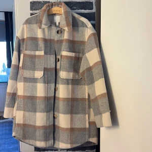 Rutig overshirt i ullmix - Overshirt i beige, grått och brunt rutmönster med stora fickor framtill och knappar längs hela framsidan. Jackan har klassisk krage och är tillverkad i en mjuk ullblandning. Perfekt för lager-på-lager och en chill look. OBS! STOR I STORLEK