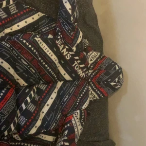 Tommy Jeans TJM Down AOP Puffer Jacket - Condition: Like new – worn 0 times, no damage Color: Navy with red lining, “New York 1985” design  Rare and bold puffer from Tommy Jeans with all-over graphic print. Super warm and stylish, perfect for winter or streetwear looks.  Filling: 60% duck down, 40% feathers  Originally bought for around 1,800 kr – selling because I never wore it.                                           Tommy Jeans TJM Down AOP Puffer Jacket (DM0DM08756)