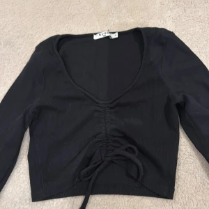Svart ribbad croppad topp NA-KD - Svart croppad topp från NA-KD med långa ärmar och djup v-ringning. Toppen har ribbad struktur och snygg snörning framtill som ger en cool detalj. Perfekt att styla med höga jeans eller kjol för en trendig look.