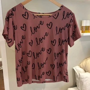 Rosa t-shirt med hjärtan och text XS - Nästa aldrig använt. Byxorna är i storlek xs men dom är stretchiga. Superfin rosa t-shirt med svarta hjärtan och texten 'love' tryckt över hela. T-shirten har rund halsringning och korta ärmar. Perfekt för dig som gillar gulliga och lekfulla prints. 🥰🥰