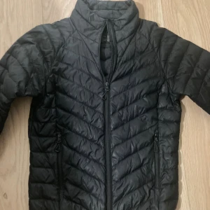 Svart pufferjacka från Neomondo - Svart pufferjacka från Neomondo i storlek S. Jackan har snyggt quiltat mönster, hög krage och dragkedja framtill samt två dragkedjeförsedda fickor. Perfekt för kyliga dagar och enkel att matcha med allt. Lätt och smidig att bära med sig.