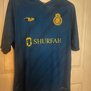 Al Nassr Ronaldo 7 blå matchtröja - Snygg blå Al Nassr fotbollströja med gula detaljer och tryck, inklusive Ronaldo 7 på ryggen. Tröjan har klubbmärke och sponsorlogga framtill, samt korta ärmar. Tillverkad i lätt och ventilerande funktionsmaterial, perfekt för fotboll eller träning.