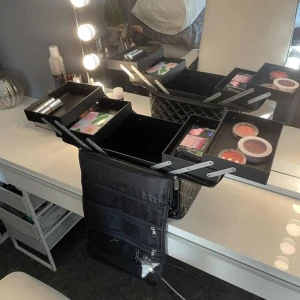 Shelas - Svart quiltad sminkväska med dragkedja - Helt oanvänd. Stor svart sminkväska med quiltad yta och flera smarta fack. Väskan har utdragbara hyllor, meshfickor med dragkedja och bärhandtag. Perfekt för att organisera smink och tillbehör. Tillverkad i glansigt syntetmaterial för en snygg look.