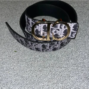 Svart grå bälte äkta D.I.O.R - Snyggt bälte från  med svart och grått mönster och stor guldfärgad CD-logga som spänne. Bältet har ett brett band och är tillverkat i skinn med tryckt logomönster över hela ytan. Perfekt accessoar för att lyfta din outfit.