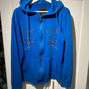Blå hoodie från Blinglucid med strass - Blå hoodie från Blinglucid i storlek M med huva och dragkedja. Framsidan har strassdetaljer med texten BLING LUCID och två änglar. Tröjan har långa ärmar, fickor och ribbade muddar. Perfekt för dig som gillar statement-plagg och coola detaljer.