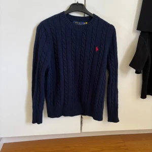 Mörkblå kabelsrickad tröja från Polo Ralph Lauren - Kabelstickad mörkblå tröja från Polo Ralph Lauren med rund halsringning och röd broderad logga på bröstet. Klassisk modell med ribbade muddar vid ärmslut och nederkant. Perfekt för lager-på-lager-stil.