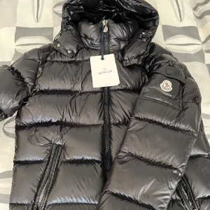 Svart Moncler pufferjacka med huva - Säljer en svart pufferjacka från Moncler med glansig finish och huva. Jackan har Moncler-logga på ärmen, dragkedja framtill och två sidofickor med dragkedja. Insidan har ett coolt serietidningsmotiv. Perfekt för dig som vill ha en snygg och varm jacka med streetkänsla.