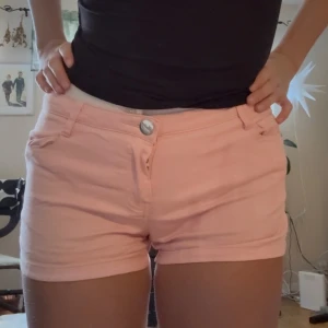 Ljusrosa lågmidjade shorts - Snygga ljusrosa shorts med klassisk femficksdesign och knappstängning fram. Modellen är kort och har bälteshällor, perfekt för sommarens varma dagar. Materialet känns mjukt och lätt, troligen bomull eller en bomullsblandning. Passformen är normal och midjan är midwaist.