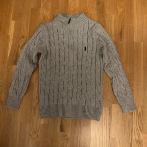 Grå kabelstickad tröja Polo Ralph Lauren - Kabelstickad grå tröja från Polo Ralph Lauren med halv dragkedja och broderad logga på bröstet. Tröjan har ribbade muddar och hög krage, perfekt för kyliga dagar. Klassisk och stilren design som funkar till allt.