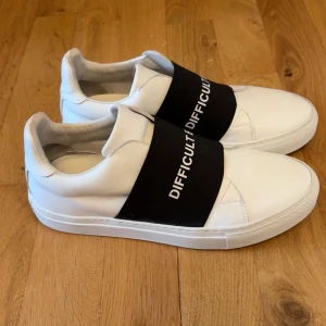 Vita sneakers Difficult by P. - Snygga vita sneakers från Difficult by P. med bred svart resår över vristen och vit text. Skorna har en stilren design med rund tå och platt sula. Tillverkade i skinn för en clean look och bekväm känsla. Perfekta för dig som gillar minimalistisk streetstyle.