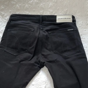 Svarta jeans från Calvin Klein - Svarta jeans från Calvin Klein Jeans med klassisk femficksdesign och diskret logga på bakfickan. Byxorna har raka ben och dragkedja i gylfen. Snygg, stilren look som funkar till det mesta. Materialet är jeans med en mjuk känsla.