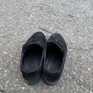 Svarta sneakers från Axel Arigato - Svarta sneakers från Axel Arigato i mocka med diskret logga på sidan. Skorna har en låg profil, platt sula och elastisk panel över vristen istället för klassisk snörning. Perfekt för dig som gillar minimalistisk och stilren design.