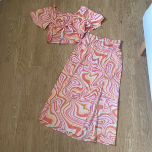 Färgglad långkjol från Lindex - Säljer en superfin långkjol från Lindex med färgstarkt psykedeliskt mönster i rosa, orange, vitt och gult. Kjolen har en rak passform och är tillverkad i ett mjukt, lätt material som känns skönt mot huden. Perfekt för dig som vill sticka ut med en cool och unik stil!