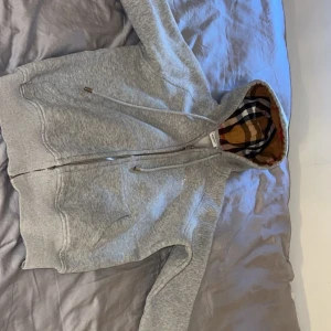 Grå Burberry hoodie med rutig huva - Säljer en grå hoodie från Burberry med klassiskt broderat emblem på bröstet och dragkedja framtill. Huvan har ikoniskt rutigt foder i beige, svart, rött och gult. Tröjan har snörning i huvan och ribbade muddar. Perfekt för dig som vill ha en stilren och lyxig look.