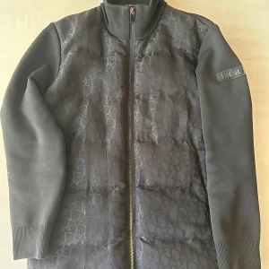 Svart pufferjacka från Dior - Säljer en svart pufferjacka från Dior med diskret mönster på framsidan och ribbade muddar. Jackan har hel dragkedja och en snygg Dior-logga på ärmen. Materialet är mjukt och känns lyxigt, perfekt för dig som vill ha något stilrent och exklusivt.
