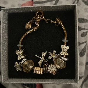 Charmarmband med fjärilar och blommor - Unikt armband i guld- och silverfärg med flera berlocker som fjärilar, blommor, trollslända och stora glittriga pärlor. Armbandet har detaljer i vitt och guld och är perfekt för dig som gillar naturinspirerade smycken med mycket detaljer.