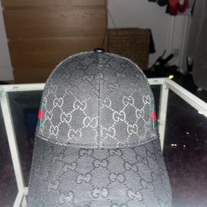 Svart keps med monogram - Svart keps -monogram över hela ytan. Sidorna har gröna och röda ränder. Justerbar rem baktill. Tillverkad i bomull och polyester, perfekt för dig som vill sticka ut.