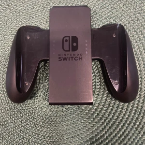 Nintendo Switch Joy-Con Grip och Joy-Con Remmar - Nintendo Switch Joy-Con Grip och två Joy-Con remmar i svart. Gripen har synliga repor och lätt slitage, men är fullt funktionell. Perfekt för att samla dina Joy-Cons till en traditionell handkontroll. Remmarna är i gott skick och ger extra säkerhet vid spel.