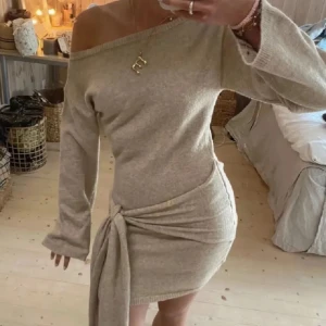 Beige stickad kortklänning med knyt - Superfin beige stickad klänning med långärmade och en stor knyt-detalj i midjan som ger en cool och trendig look. Klänningen är kort och har en mjuk, cozy känsla som passar perfekt till boots. Perfekt för dig som gillar stilrena och moderna outfits.