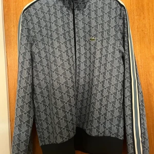 Mönstrad grå ziptröja från Lacoste storlek L - Säljer en grå långärmad ziptröja från Lacoste med vitt geometriskt mönster och klassisk krokodillogga på bröstet. Tröjan har svarta muddar, dragkedja framtill och vita ränder längs ärmarna. Regular fit och perfekt för en sportig look. Mycket fint skick, använd vid 2 tillfällen.