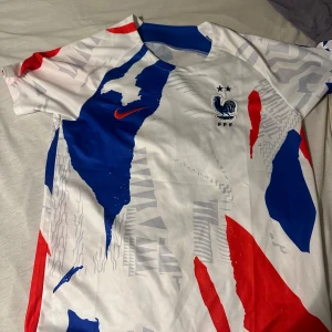 Frankrike Nike fotbollströja blå/röd/vit - Snygg Frankrike fotbollströja från Nike med FFF-logga och två stjärnor på bröstet. Tröjan har korta ärmar och är designad i vitt med blå och röda grafiska detaljer. Perfekt för dig som vill visa ditt stöd för det franska landslaget.