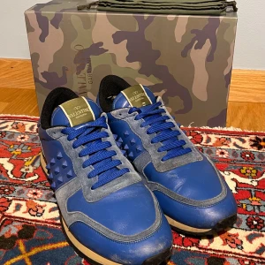 Valentino Garavani Rockrunner blå sneakers - Snygga Valentino Garavani Rockrunner sneakers i blått skinn och mocka med coola nitar på sidorna. Skorna har blå snörning, svart innersula och robust gummisula. Kommer med originalkartong i camouflage-mönster. Perfekta för dig som vill sticka ut med stil.   Skorna har mindre slitage vid hälen och lådan är i acceptabelt skick.