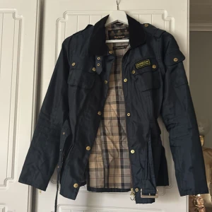 Mörkblå Barbour International jacka - Snygg mörkblå Barbour International jacka med guldiga detaljer, bälte i midjan och klassiskt rutigt foder. Jackan har flera fickor, dragkedja och knappar framtill samt en krage i manchester. Perfekt för dig som gillar stilren och tidlös design. Passar storlek XS/S