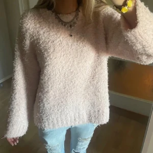 Ljusrosa fluffig stickad tröja - Supermysig ljusrosa stickad tröja med fluffig textur och rund hals. Tröjan har en loose passform och långa ärmar, perfekt för kyliga dagar. Ribbstickade muddar vid ärmslut och nederkant ger en extra snygg detalj.