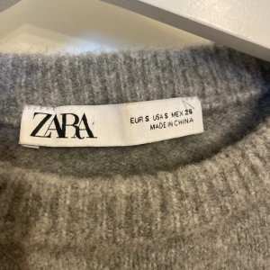 Grå stickad tröja från Zara - Mysig grå stickad tröja från Zara med korta ärmar och rund halsringning. Tröjan har en enkel och stilren design med ribbade muddar vid ärmslut och nederkant. Perfekt för lager-på-lager och passar till allt.
