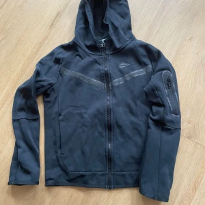 Svart hoodie plus byxor x2 från Nike - Svart hoodie från Nike i stl M 137-147cm (junior) med hel dragkedja framtill och huva. Jackan har en diskret logga på bröstet, fickor i sidorna och en extra ficka med dragkedja på ena ärmen.                                                                       Två stycken tillhörande byxor i svart i stl L 147-158 (junior) i bra skick. Lite synlig användning på knäna. Byxorna har fickor på båda sidor samt en ficka med dragkedja.
