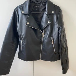 Svart skinnjacka från The Black Label - Snygg svart skinnjacka från The Black Label Collection i bikerstil. Jackan har silvriga dragkedjor, tryckknappar och klassisk krage. Materialet är syntetiskt skinn och modellen är kortare med lång ärm och detaljerade sömmar. Perfekt för en edgy look.