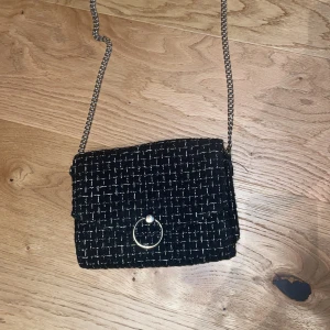 Svart handväska med kedjerem - Stilren svart handväska med rutigt mönster i vitt och guldtråd. Väskan har en snygg kedjerem med läderparti för axeln. Från H&M, köpte för runt 200 för något år sedan❣️