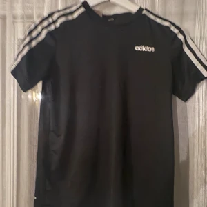 Svart Adidas t-shirt med ränder - Svart t-shirt från Adidas med klassiska vita ränder längs axlarna och ärmarna. Adidas-logga i vitt på bröstet. T-shirten har rund hals och är tillverkad i mjuk bomull. Perfekt för en sportig och avslappnad stil.