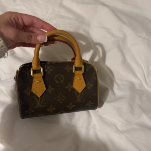 Louis Vuitton monogram handväska - Superfin liten handväska från Louis Vuitton i klassiskt monogrammönster. Väskan är brun med ljusbruna handtag och detaljer i skinn, samt guldfärgade metalldetaljer. Stängs med dragkedja och har en kompakt, boxig form som är perfekt för det viktigaste.