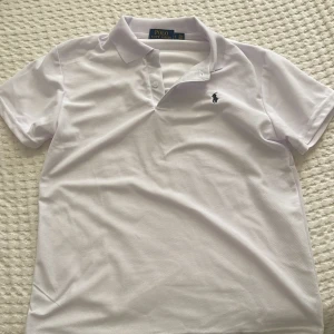 Vit polo t-shirt från Ralph Lauren - Vit klassisk polo t-shirt från Ralph Lauren med korta ärmar och krage. T-shirten har den ikoniska broderade loggan på bröstet och är gjord i mjuk bomull. Perfekt för dig som gillar stilrena och tidlösa plagg.