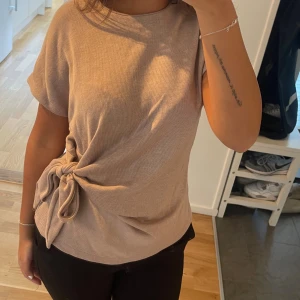 Beige topp med knyt i sidan - Säljer en beige topp med korta ärmar och rund hals. Toppen har en snygg knytning i sidan som ger en cool och unik look. Materialet känns mjukt och har en lätt stickad struktur. Perfekt för dig som gillar enkla men stilrena detaljer.