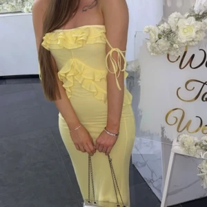 Gul långklänning med volanger - Säljer en ljusgul långklänning med offshoulder och snygga knytband vid armarna. Klänningen har flera volangdetaljer framtill som ger en cool och romantisk vibe. Passformen är tight och materialet känns mjukt och stretchigt, perfekt för dig som vill sticka ut. Köpt från asos.