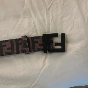 Brunt Fendi bälte med FF-logga - Säljer ett brunt bälte från Fendi med ikoniskt FF-mönster i svart och en stor svart metallspänne formad som Fendi-loggan. Bältet är gjort i skinn och har en bred, stilren design som sticker ut.
